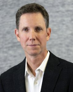Professional headshot of a middle-aged man in a dark blazer and white shirt against a gray backdrop.