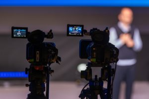 Two professional video cameras on tripods recording a speaker on a blue-lit stage in the background.